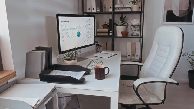 No people shot of cozy workspace with computer monitor with business document on screen, coffee mug on desk, white leather rolling chair and paintings on wall in modern office with biophilic design