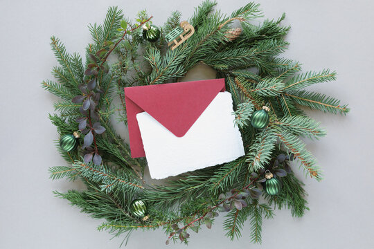 Overhead view of an envelope and blank card in the middle of a coniferous festive Christmas wreath decorated with christmas baubles on a grey background