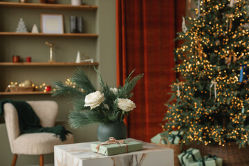 Vase of white roses and conifer branches on a table and a wrapped Christmas gift in front of an illuminated Christmas tree in a living room decorated for the holidays
