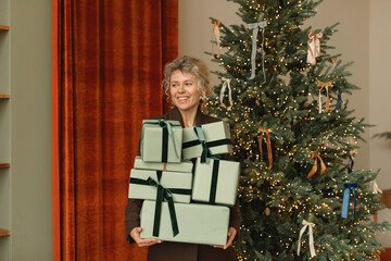 Smiling woman standing next to an illuminated Christmas tree in a living room holding a stack of Christmas gifts