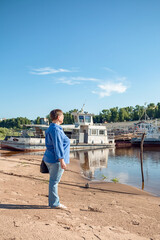 An elderly woman stands on the river bank near an old river transport.