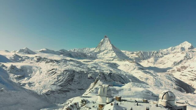 A mountain range with a snow covered peak and a building in the foreground. The sky is clear and blue