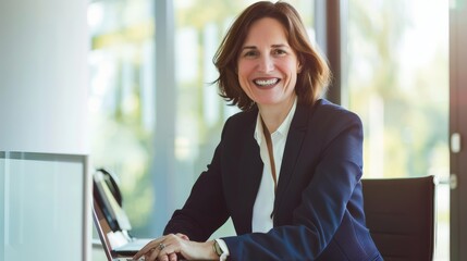 Fototapeta premium A businesswoman sitting by her laptop, dressed in a professional suit, smiling confidently with a modern office setting in the background.