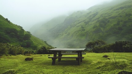 An empty wooden picnic table surrounded by lush green hills and enveloped in a light mist, suggesting a peaceful, remote outdoor setting.