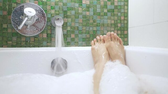 A person in a bathtub with the water running. The person is wearing a white towel and has their feet in the water