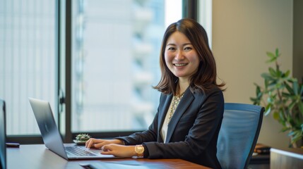 A professionally dressed woman smiling confidently while working on a laptop in a modern office environment with large windows and city view.
