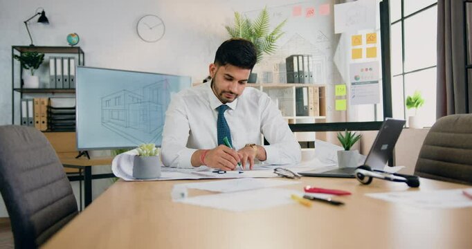Man constructor drawing on blueprint using information from computer project sitting at drafting table in engineering office