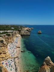 The photo shows the impressive limestone arches near Benagil Cave in the Algarve, Portugal. Crystal-clear green waters flow beneath the towering cliffs, set against a bright blue sky and rugged coastl