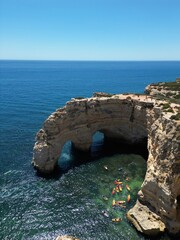 The photo shows the impressive limestone arches near Benagil Cave in the Algarve, Portugal. Crystal-clear green waters flow beneath the towering cliffs, set against a bright blue sky and coastline.