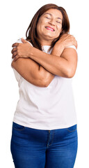 Middle age latin woman wearing casual white tshirt hugging oneself happy and positive, smiling...