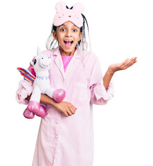 Cute african american girl wearing sleep mask and pajama holding pink teddy unicorn celebrating victory with happy smile and winner expression with raised hands