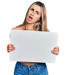 Fototapeta premium Hispanic young woman holding blank empty banner in shock face, looking skeptical and sarcastic, surprised with open mouth