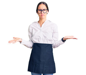 Beautiful brunette young woman wearing professional waitress apron clueless and confused with open arms, no idea concept.
