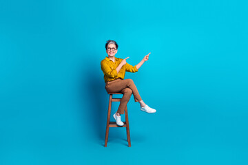 Full length photo of charming positive woman wear blue blouse eyeglasses sitting chair pointing empty space isolated blue color background