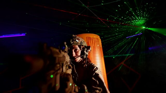 A woman in a military uniform with a submachine gun, illuminated by green and purple lasers, prepares for action in a dark room. The atmosphere is dynamic and intense.