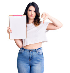 Beautiful young brunette woman holding clipboard with blank space with angry face, negative sign showing dislike with thumbs down, rejection concept