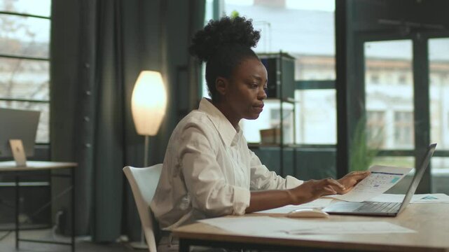 Young African American business woman sitting on chair use laptop hold paper working in office. Look serious. Camera moving around. 360 degrees