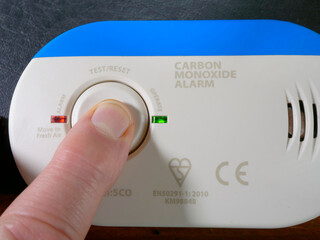 Closeup of a finger tip pressing the button to test the operation of a household carbon monoxide alarm, with red and green lights flashing.