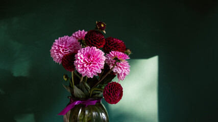 Bouquet of dahlias in green glass vase on green background with sunlight.Vase of flowers stands on wooden table.Concept: greeting card, autumn holidays.Copy space.Selective focus.