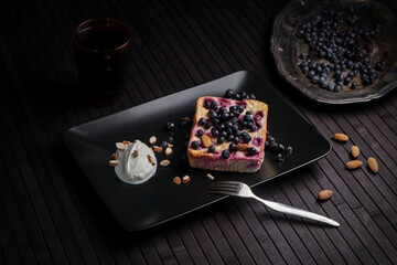 A rustic dessert featuring a slice of blueberry cake on a black plate, served with a dollop of whipped cream and almonds. Fresh blueberries and almonds scattered around enhance the dark, cozy mood