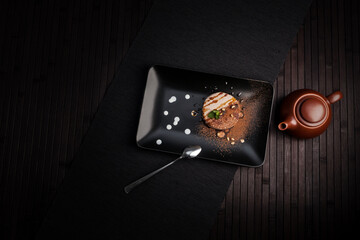 A sophisticated dessert on a black plate with artistic drizzle of sauces and dusting of cocoa, complemented by a modern teapot. The minimalistic, dark-toned setup creates an elegant atmosphere.