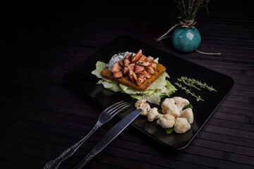 A savory dish featuring seasoned chicken over lettuce and a crispy base, served with roasted cauliflower and fresh thyme on a black plate. The elegant plating creates a refined dining experience.