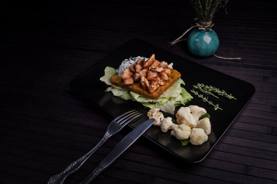A savory dish featuring seasoned chicken over lettuce and a crispy base, served with roasted cauliflower and fresh thyme on a black plate. The elegant plating creates a refined dining experience.