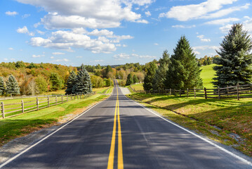 Empty road between fenced fields in a rolling forested landscape on a clear autumn day