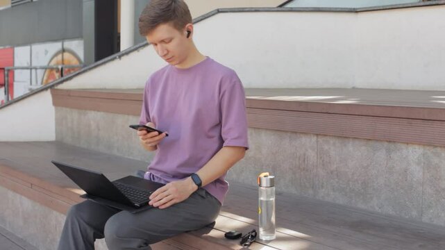 Handsome guy in casual clothing using cell phone with headphones on city street. Young man scrolling through news feed on phone.