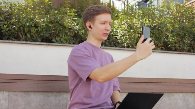 Closeup man making video call with headphones and smartphone in park, waving at people on phone screen. Online communication concept. 
