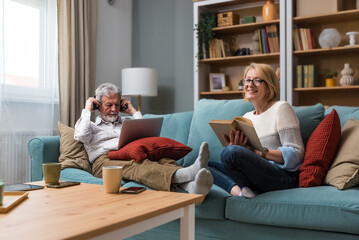 Life goals Retired senior couple man and woman enjoying their life at home, using laptop computer and reading a books, relaxing, listening music or podcast spending quality time together in retirement