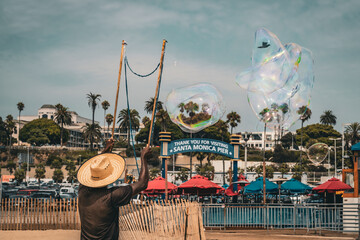 An African American man wearing a hat and making big soap bubbles on Santa Monica beach
