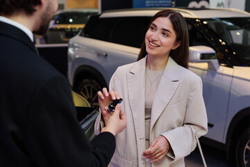Medium shot of smiling young woman in elegant beige suit talking to auto dealer taking key of new...