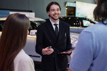 Medium shot of smiling Central Asian male auto dealer in black suit holding key of new car, while...