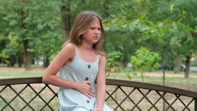 Kid girl sitting alone on the bench in the park, in pain holding belly, feeling stomach ache, symptoms or diarrhea. Upset preteen girl suffering from indigestion, flatulence and abdominal problem