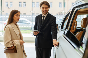 Medium full shot of smiling male auto dealer in black suit opening car door, presenting demo model...
