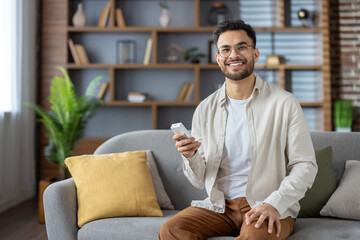 Obraz premium Smiling hispanic man relaxing at home, sitting comfortably on couch, holding air conditioner remote control, enjoying modern technology, and creating cozy atmosphere in well-furnished living room