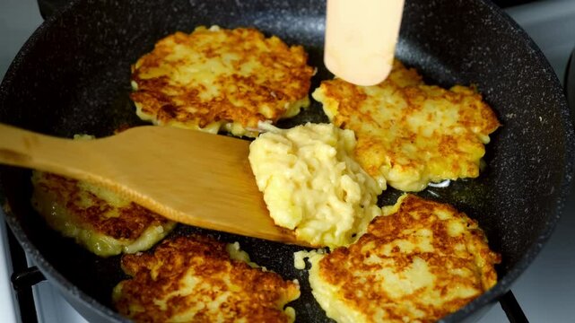 Potato pancakes, draniki cooking in frying pan on gas oven on kitchen at home. Cook turns over draniki using wooden spatula. Preparing fresh dish. Cuisine, culinary, recipe. Traditional Russian food.