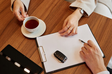 Close up on car key and hands of female client buying new vehicle and auto dealer providing her with purchase contract at dealership center