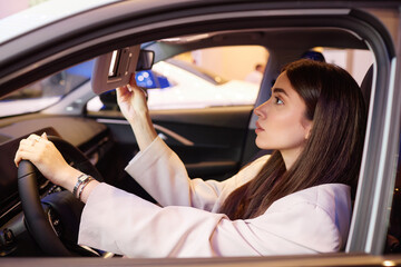 Side view of young woman behind wheel looking in visor mounted mirror while testing demo vehicle...
