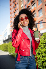 Vertical photo of pretty cheerful lady wear red leather jacket drinking beverage listen modern gadget outdoors city urban street