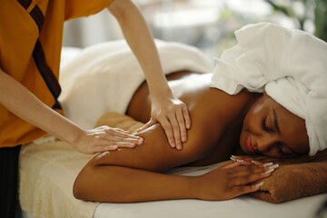 A woman is receiving relaxing massage on a professional table with soft lighting, her eyes closed, wearing a towel, and a towel covering her head while another person is massaging her arm
