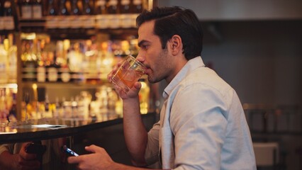 Smart man drinking cocktail in Old fashioned waiting friends or couple while chatting on smartphone to wait at counter bar with bartender preparing drinks at nightclub Friday meeting night. Vinosity.