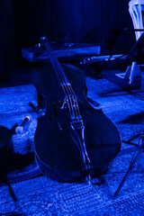 Striking cello resting on a patterned rug under blue stage lighting with a microphone and laptop nearby, evoking a serene musical setting © Olga Mazo