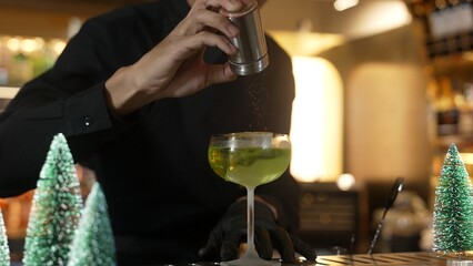 Cropped bartender garnishing caramelized citrus slice on cube ice in green Midori Sour cocktail served in stylish stemmed glass. Perfect for sophisticated bar settings christmas tree theme. Vinosity.