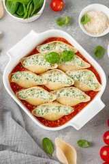 Conchiglioni pasta stuffed ricotta cheese and spinach with tomato sauce in a white baking dish on a gray concrete background.