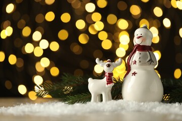 Cute decorative snowman and deer on artificial snow against blurred lights. Space for text