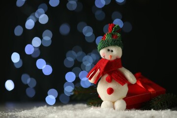 Cute decorative snowman on artificial snow against blurred lights. Space for text