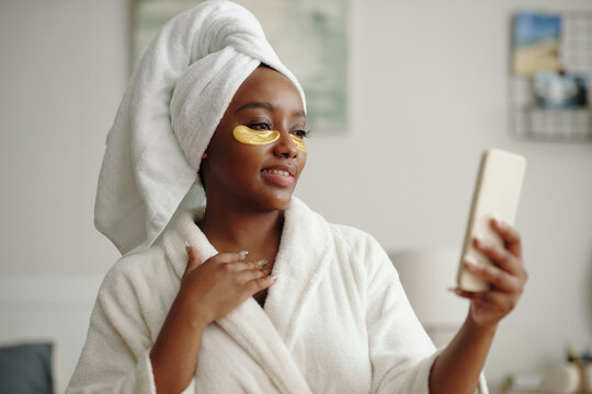 Smiling woman in white bathrobe and towel holding phone while wearing undereye patches with hand near chest, standing in softly lit room during daily skincare routine