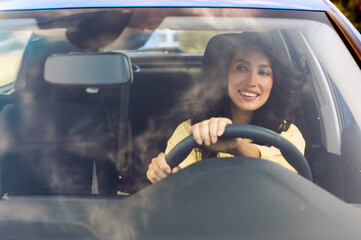Positive woman driving own modern car, holding steering wheel and smiling, looking through front...